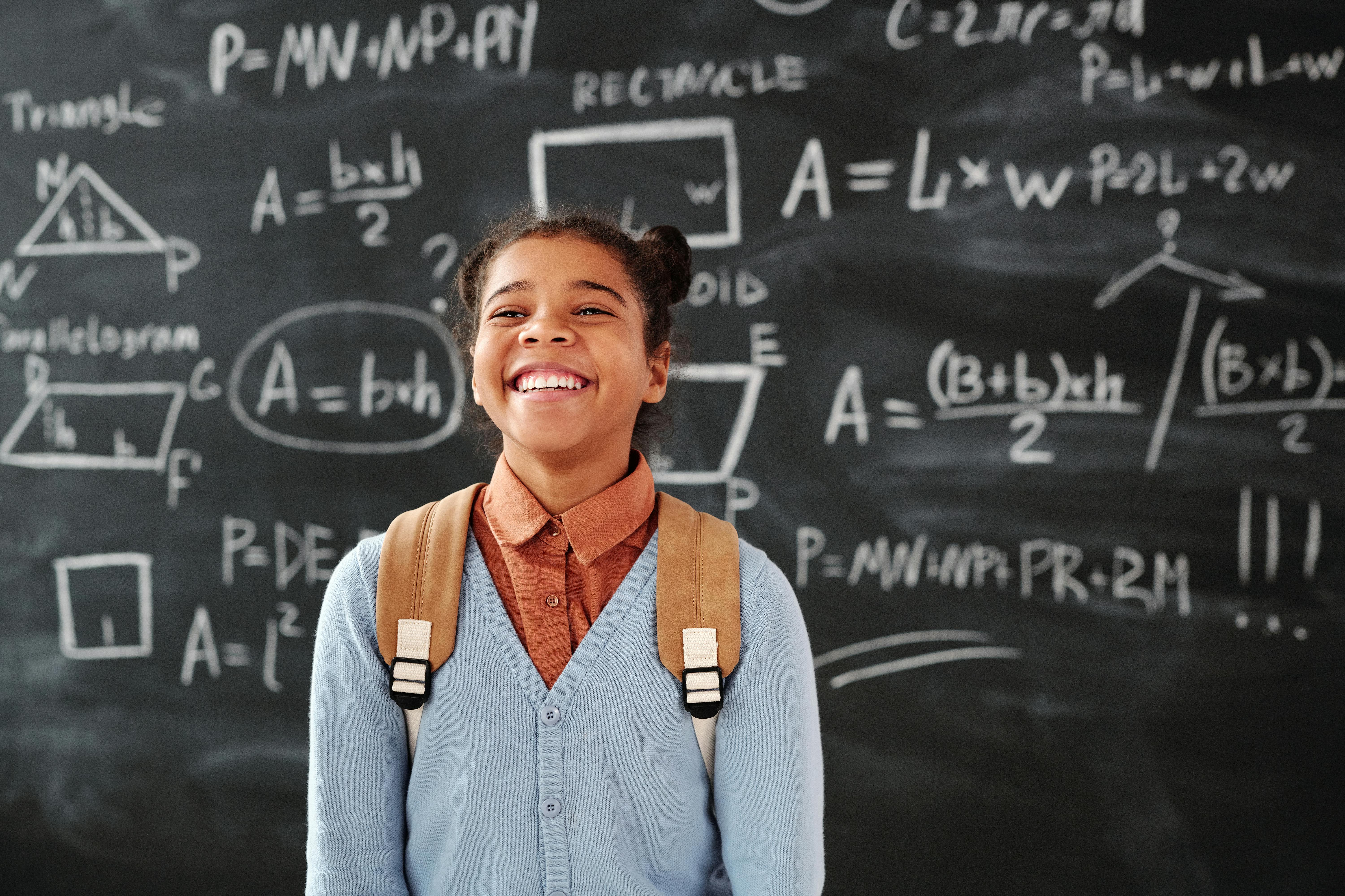 A smiling student with a backpack in front of a chalkboard covered in math