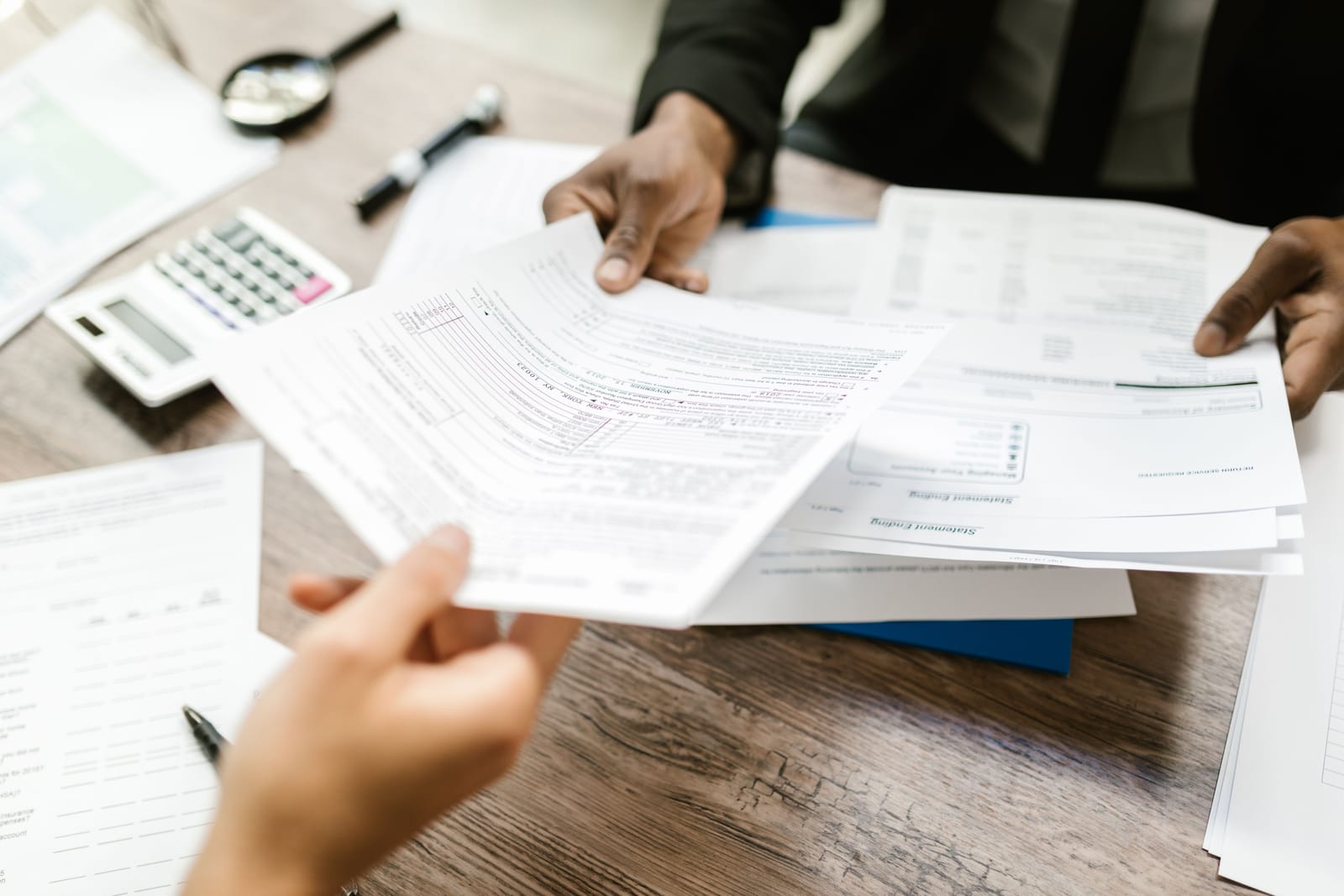 Two people passing tax and benefits paperwork across a wooden desk with a calculator and a magnifying glass