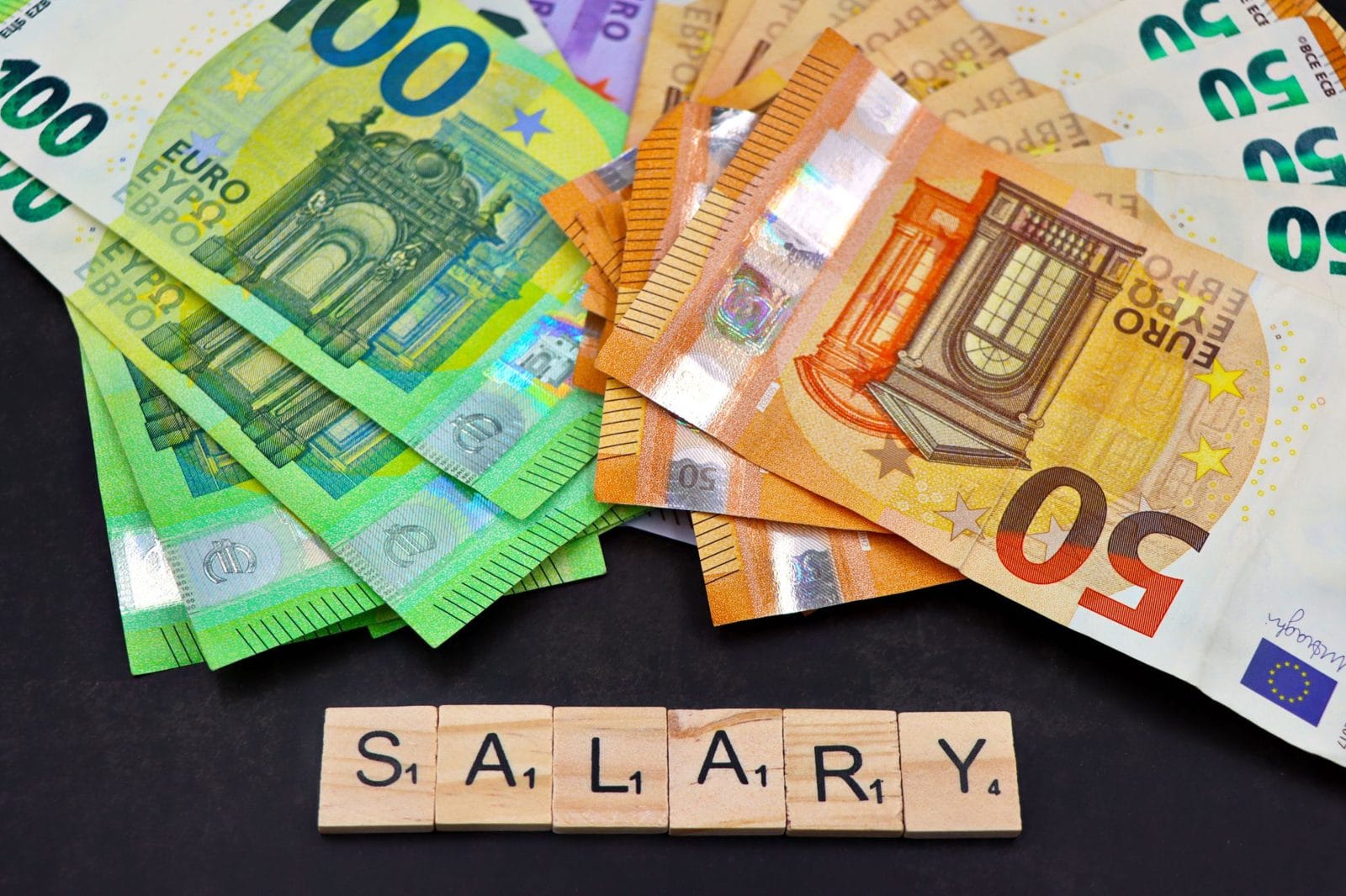 Fanned euro banknotes in green and orange above wooden letter tiles spelling the word SALARY on a dark surface